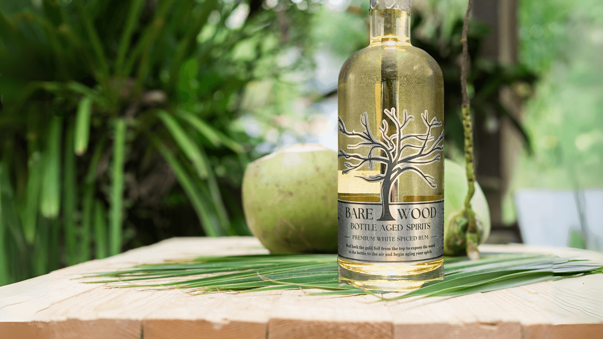 Bottle of Bare Wood bottle-aged spirits on a wooden table with coconut and greenery in the background.